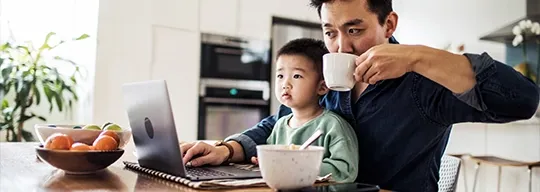 A father checks his laptop while drinking a cup of coffee with his son on his lap.